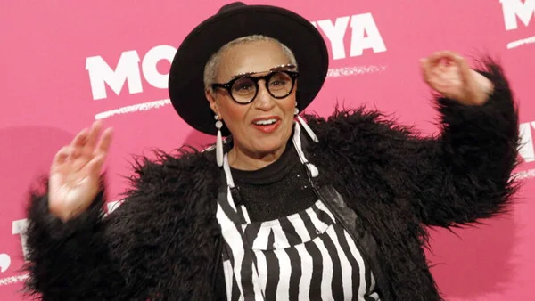 Lââm coiffée de cheveux gris et d'un chapeau noir posant avec les mains levées.