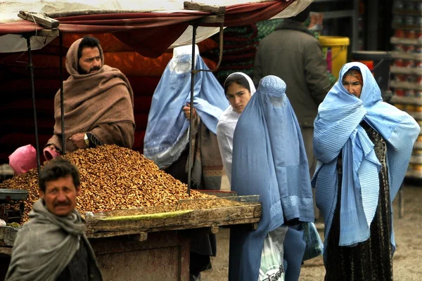 Étal de marché avec un vendeur et plusieurs femmes en burqas bleues au premier plan.