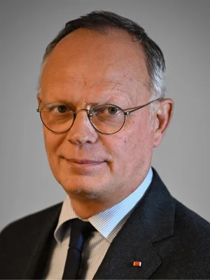 Édouard Philippe en costume sombre, chemise bleue rayée et lunettes rondes, portraituré sur fond gris uni.