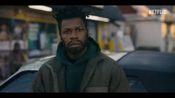 John Boyega dans une rue urbaine, portant une veste verte, dans une scène du film.