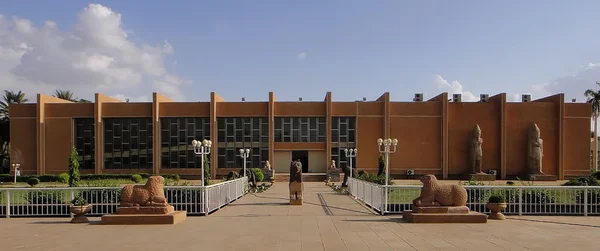 Grand bâtiment symétrique brun du musée avec de grandes fenêtres, bordé par des statues de lions et de figures humaines dans un parc paysager.