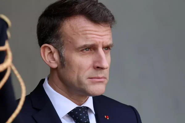 Emmanuel Macron en costume sombre debout sur fond gris.