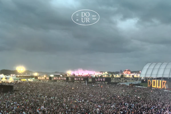 Foule réunie sur un pré devant un grand panneau blanc marquant 'DOUR', entouré d'arbres et de bâtiments.