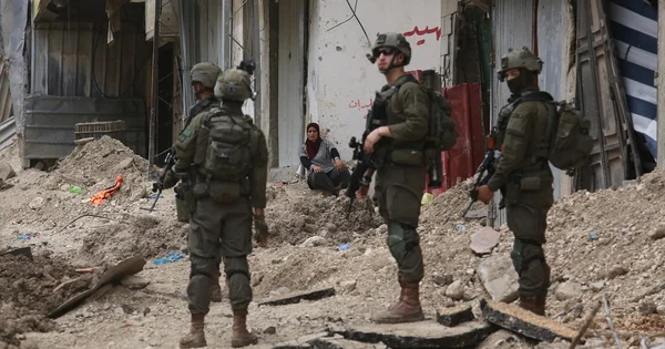 Quatre soldats en tenue militaire se tiennent sur une rue encombrée de gravats devant un bâtiment abîmé