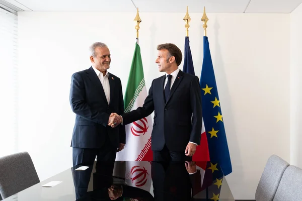 Emmanuel Macron et un homologue iranien se serrent la main devant les drapeaux français, iranien et européen.
