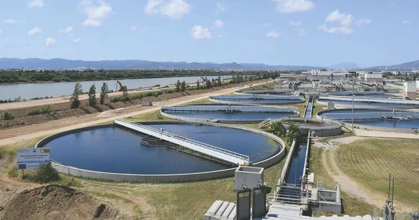 Vue d'une station d'épuration à Barcelone avec ses grands bassins circulaires, une rivière et les montagnes au loin.