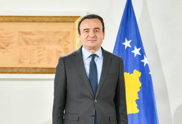 Homme en costume devant un drapeau du Kosovo avec étoiles blanches et carte jaune en arrière-plan.