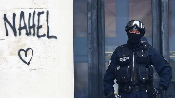 Policier en tenue tactique devant un mur aux graffitis 'NAHEL'.