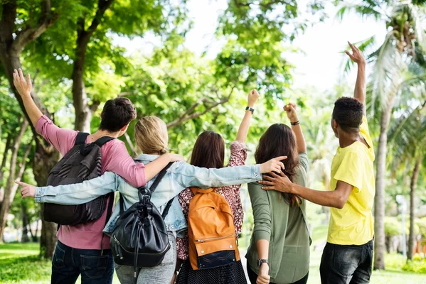 Cinq jeunes de dos lèvent les bras en signe de joie au milieu d'un parc avec des arbres verts.