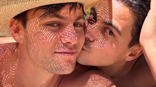 Deux hommes torse nu, dont l'un porte un chapeau de paille, s'embrassant.