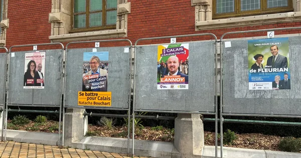 Plusieurs affiches électorales installées sur une grille métallique face à une façade en briques.