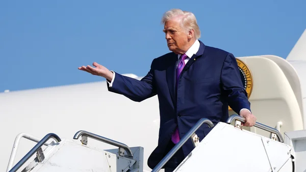 Un homme en costume bleu marine et cravate violette se tient au sommet des escaliers d'un avion, la main tendue.