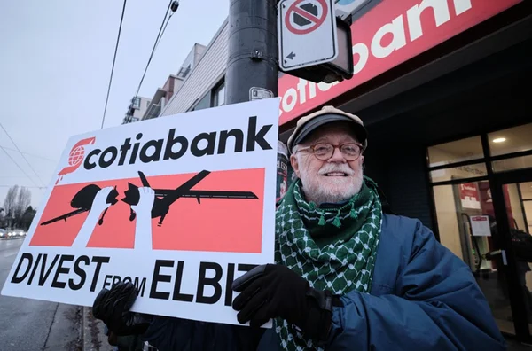 Un homme barbu porte une pancarte appelant au désinvestissement d'Elbit devant une agence Scotiabank