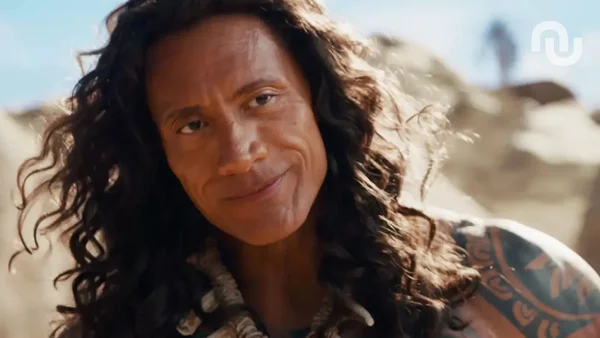Dwayne Johnson en Maui avec sa chevelure bouclée et un vêtement à col en fourrure, devant un décor rocheux.