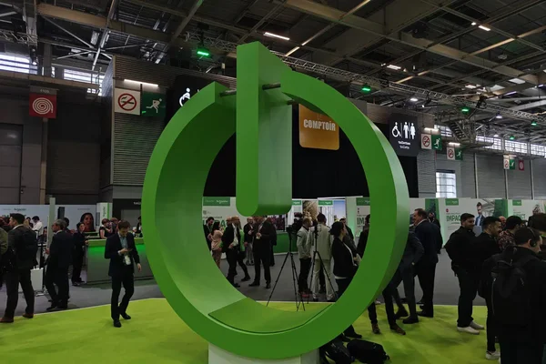 Sculpture verte en forme de bouton d'alimentation sur un socle lumineux lors d'un salon professionnel animé, entourée de participants en tenue d'affaires.