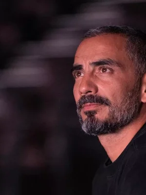 Portrait rapproché de Julien Mairesse, chemise noire, cheveux et barbe grisonnants, sur fond sombre.