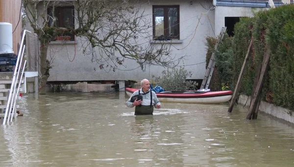 Un homme âgé debout dans l'eau devant sa maison, avec un bateau à proximité.