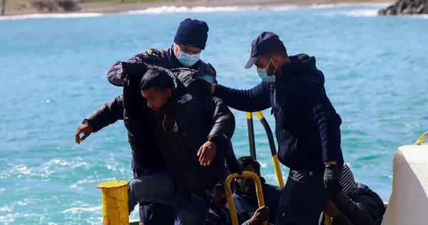Un jeune migrant assisté par deux personnels en uniforme lors de son débarquement sur un quai, eau turquoise en arrière-plan.