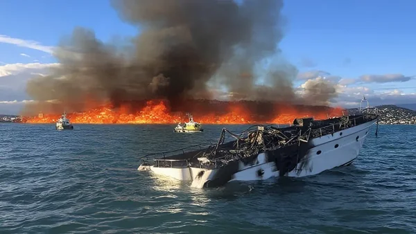 Un bateau blanc détruit flotte sur l'eau près d'un incendie sur la côte.