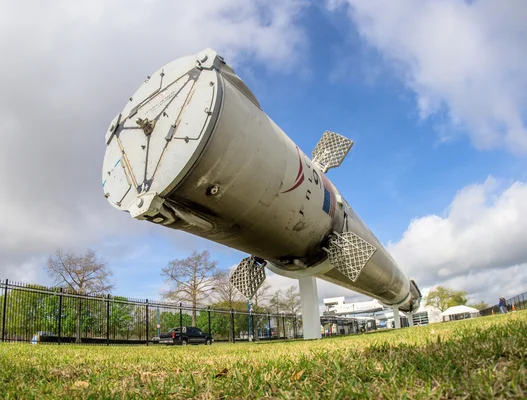 Un gros composant cylindrique d'une fusée, probablement un booster, est exposé sur une herbe près d'une clôture.