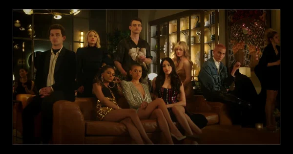 Groupe de personnages réunis dans un intérieur élégant, certains assis sur un canapé marron.