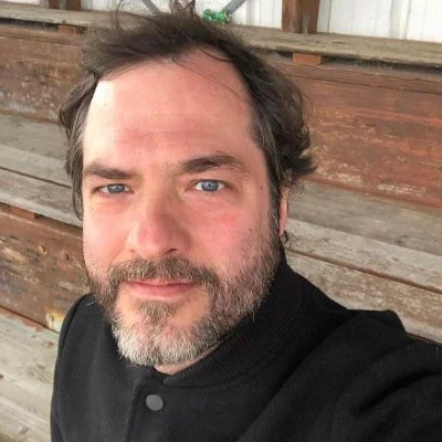 Portrait d'un homme en veste noire posant devant un fond en planches de bois.