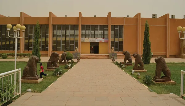Bâtiment moderne orange avec grandes fenêtres et allée pavée bordée de statues d'animaux en pierre, avec un enfant en chemise rouge marchant à gauche.