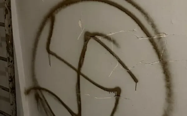 Une croix gammée peinte en brun sur un mur fissuré, près d'une échelle métallique.