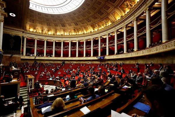 Intérieur de l'hémicycle de l'Assemblée nationale avec ses sièges rouges et son architecture ornée, durant une session parlementaire.
