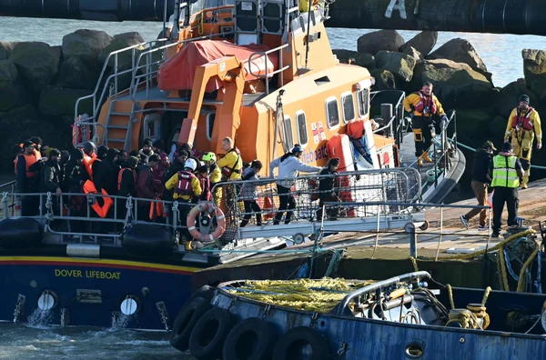 Des migrants en gilets de sauvetage débarquant d'un canot de sauvetage près d'un rivage rocheux, assistés par des sauveteurs.