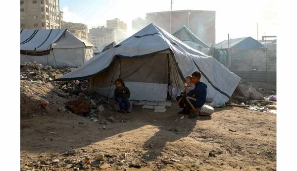 Campement de tentes de fortune au milieu des ruines, avec un homme tenant un enfant.