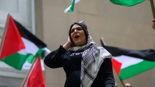 Une personne en hijab bleu clair et keffiyeh devant un drapeau palestinien dans un décor urbain.