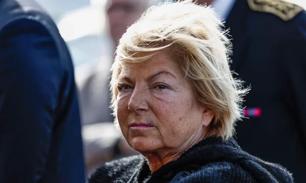 Visage centré d'une femme en vêtement foncé, avec des personnes indistinctes en arrière-plan.