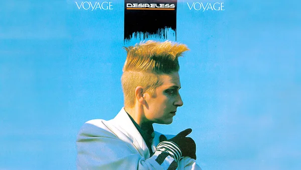 Desireless avec sa coiffure emblématique et une veste blanche devant un fond bleu.