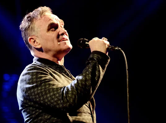 Le chanteur Morrissey chantant sur scène, portant une chemise à motifs sur fond sombre.