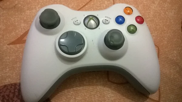 Une manette Xbox 360 blanche, identifiable par ses joysticks gris et ses boutons colorés, reposant sur un tissu à motifs.