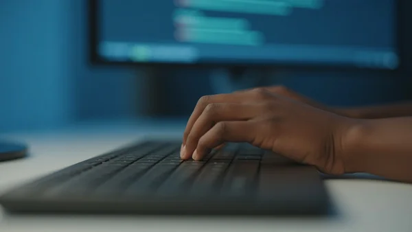 Vue en gros plan des mains d'une personne qui tapent sur un clavier d'ordinateur, écran de portable flou en arrière-plan affichant du code et des graphiques, lumière bleue de bureau