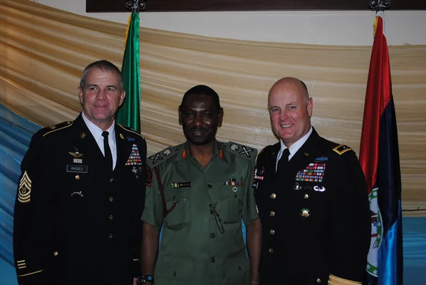 Une personne en uniforme militaire vert et casquette noire se tient dans une rue avec des structures en bois de chaque côté.
