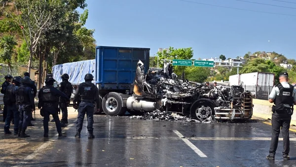 Groupe de policiers en uniforme stationnant près d'un camion lourdement calciné sur une route mouillée sous un ciel dégagé.