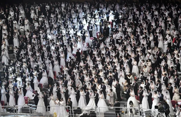 Un immense rassemblement de couples en robes de mariage blanches et costumes noirs, alignés dans une grande salle.