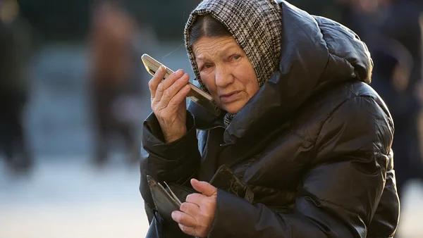 Une personne âgée tenant un téléphone à l'oreille et un portefeuille en main, debout à l'extérieur au milieu d'autres passants.