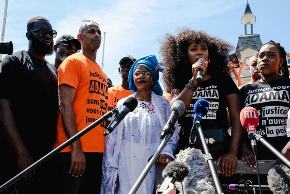 Point presse du Comité Adama avec micros de médias, militants en tee-shirts noirs et femme en tenue blanche au premier plan.