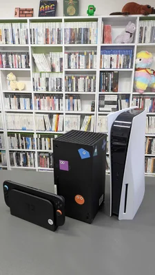 Une PlayStation 5 blanche parmi les trois consoles de génération actuelle posées sur une table devant une bibliothèque de jeux.