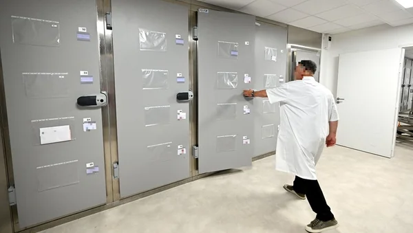 Un homme en blouse blanche devant des unités de stockage funéraire dans une salle blanche et carrelée d'un établissement hospitalier.