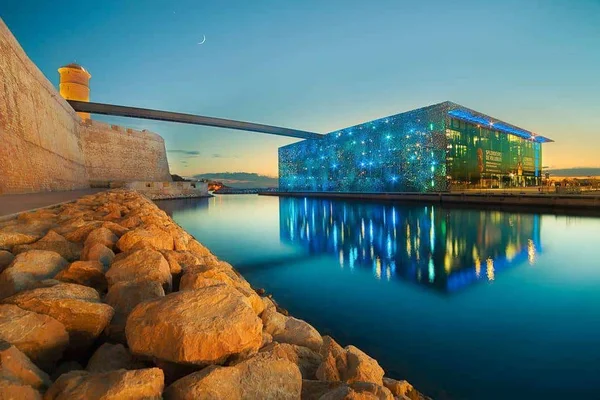 Le Mucem illuminé en bleu au crépuscule avec le reflet dans l'eau et un croissant de lune.