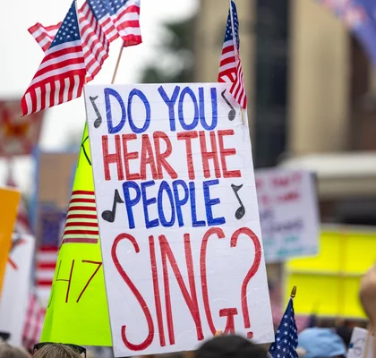 Pancarte « DO YOU HEAR THE PEOPLE SING?» lors d'une manifestation, entourée de drapeaux américains.