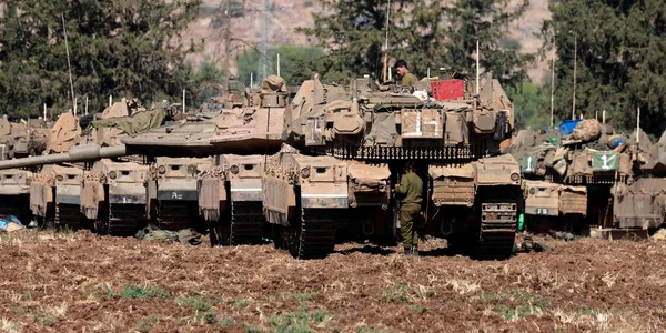 Des chars de l'armée israélienne déployés dans un champ avec des soldats en uniforme sur et autour des véhicules, sur un terrain rocailleux.