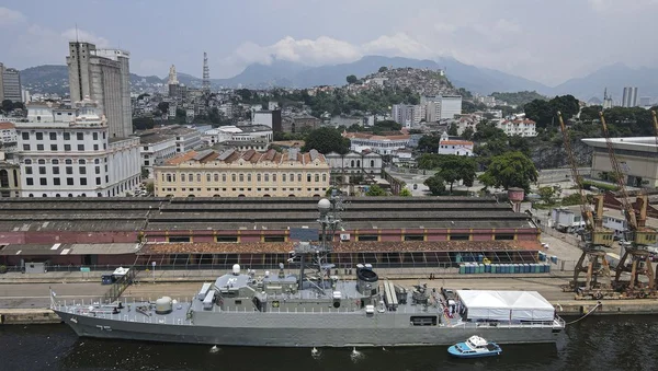 L'IRIS Dena amarré dans un port avec un paysage urbain en arrière-plan, dont un bâtiment jaune au toit rouge.