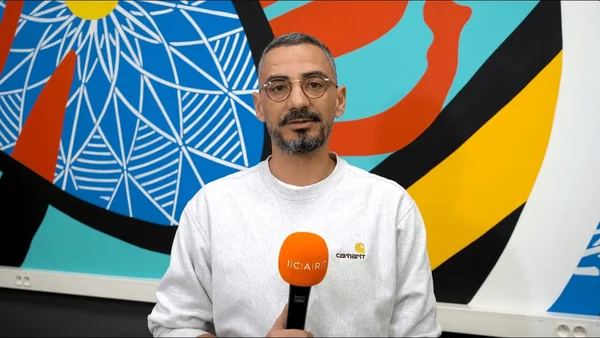Julien Mairesse avec un micro orange devant une fresque abstraite colorée, en sweat Carhartt gris clair et lunettes.