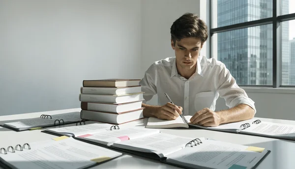 Vue en contre-plongée d'un candidat sérieux assis à un bureau lumineux, une pile de livres et de dossiers juridiques ouverts devant lui, prenant des notes sur un cahier à spirale